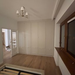 Stolarz Łukasz Kołodziejczyk - Jasne, matowe szafy wnękowe od podłogi do sufitu z małymi, złotymi uchwytami, widok na drewnianą podłogę i częściowo złożone łóżko w pokoju w trakcie remontu.