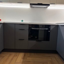 Stolarz Łukasz Kołodziejczyk - Szare, matowe fronty szafek kuchennych z poziomymi uchwytami, piekarnik w folii ochronnej, parkiet ułożony w jodełkę.