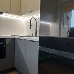 Stolarz Łukasz Kołodziejczyk - Nowoczesna kuchnia: szare szafki dolne z czarnymi uchwytami, beżowe szafki górne z podświetleniem LED, czarna bateria kuchenna, drewniana podłoga.