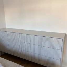 Stolarz Łukasz Kołodziejczyk - Jasnoszara komoda na metalowych nóżkach z ryflowanymi frontami szuflad, stojąca na drewnianej podłodze przy białej ścianie w sypialni.