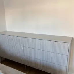 Jasnoszara komoda na metalowych nóżkach z ryflowanymi frontami szuflad, stojąca na drewnianej podłodze przy białej ścianie w sypialni.