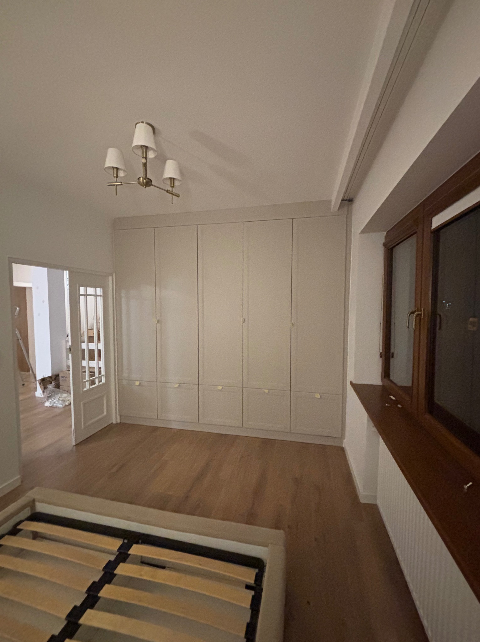 Jasne, matowe szafy wnękowe od podłogi do sufitu z małymi, złotymi uchwytami, widok na drewnianą podłogę i częściowo złożone łóżko w pokoju w trakcie remontu.