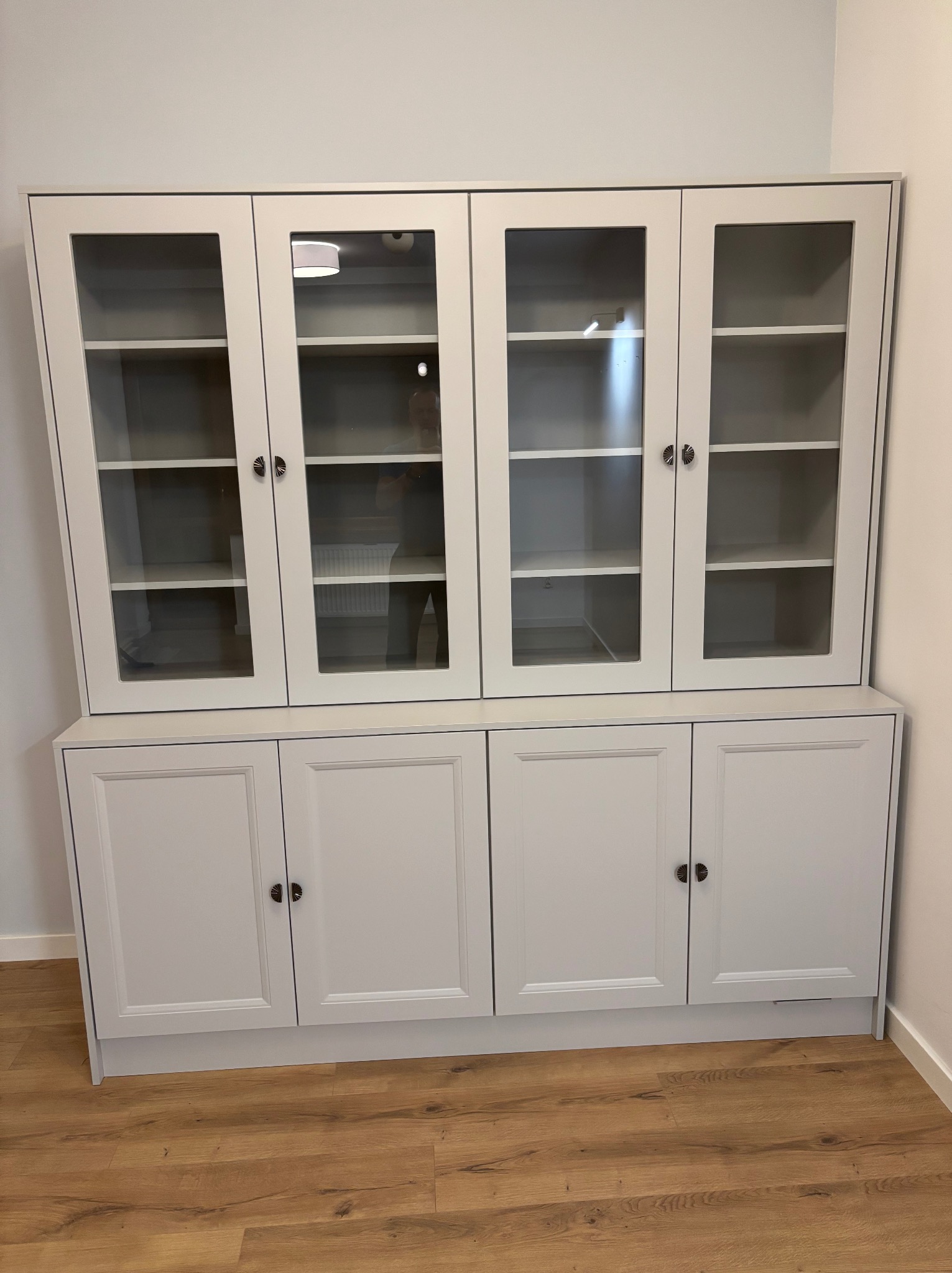 Jasnoszara witryna z przeszklonymi górnymi szafkami i pełnymi dolnymi, z ozdobnymi uchwytami, na drewnianej podłodze.