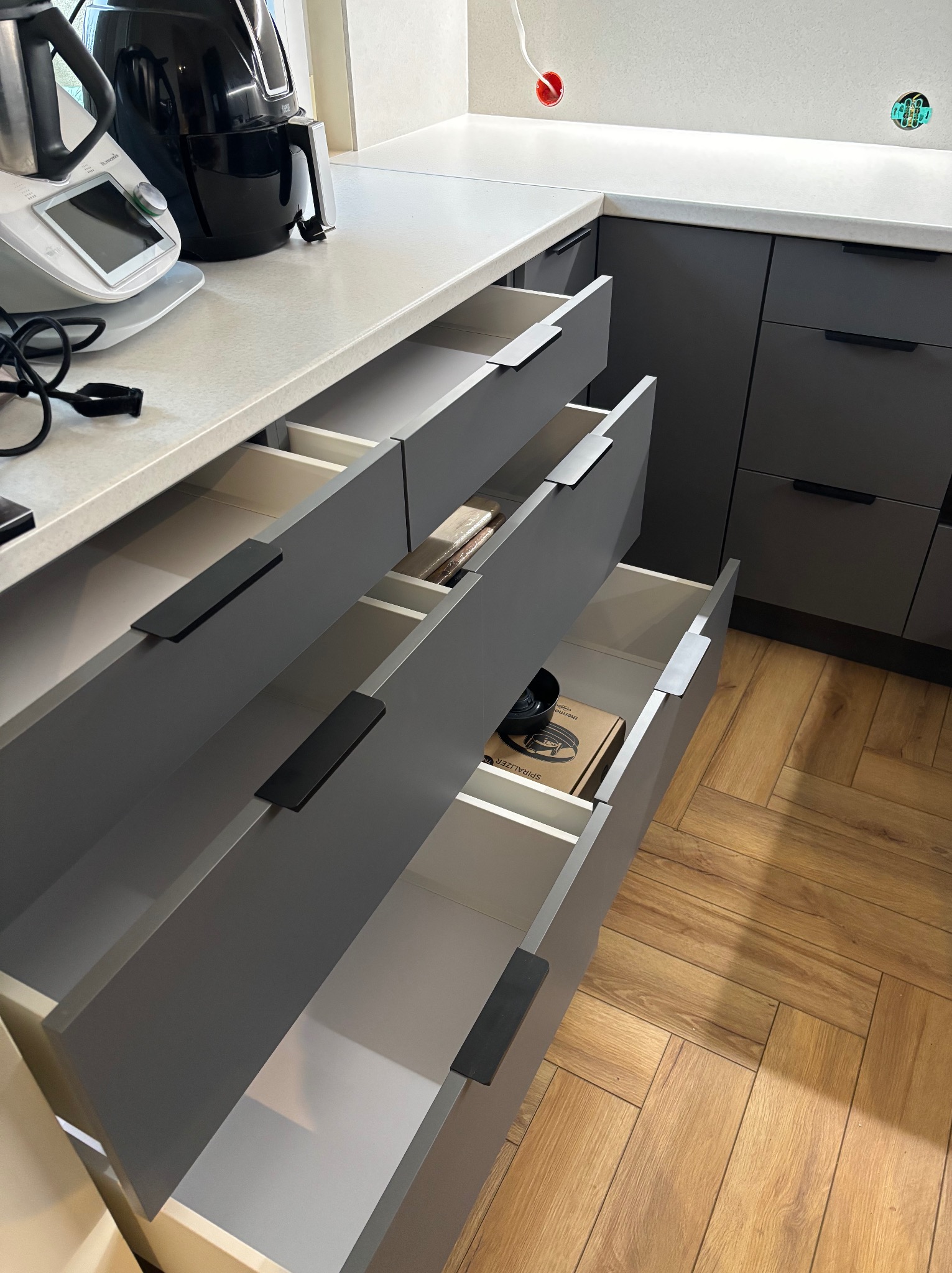 Szare szafki kuchenne z otwartymi szufladami, widoczny sprzęt AGD na blacie i drewniana podłoga w jodełkę.