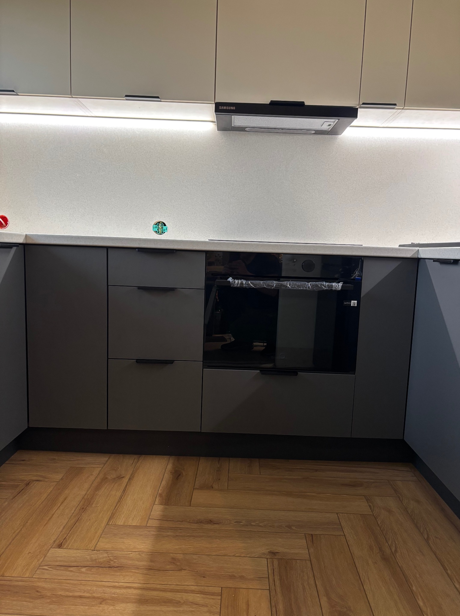 Szare, matowe fronty szafek kuchennych z poziomymi uchwytami, piekarnik w folii ochronnej, parkiet ułożony w jodełkę.