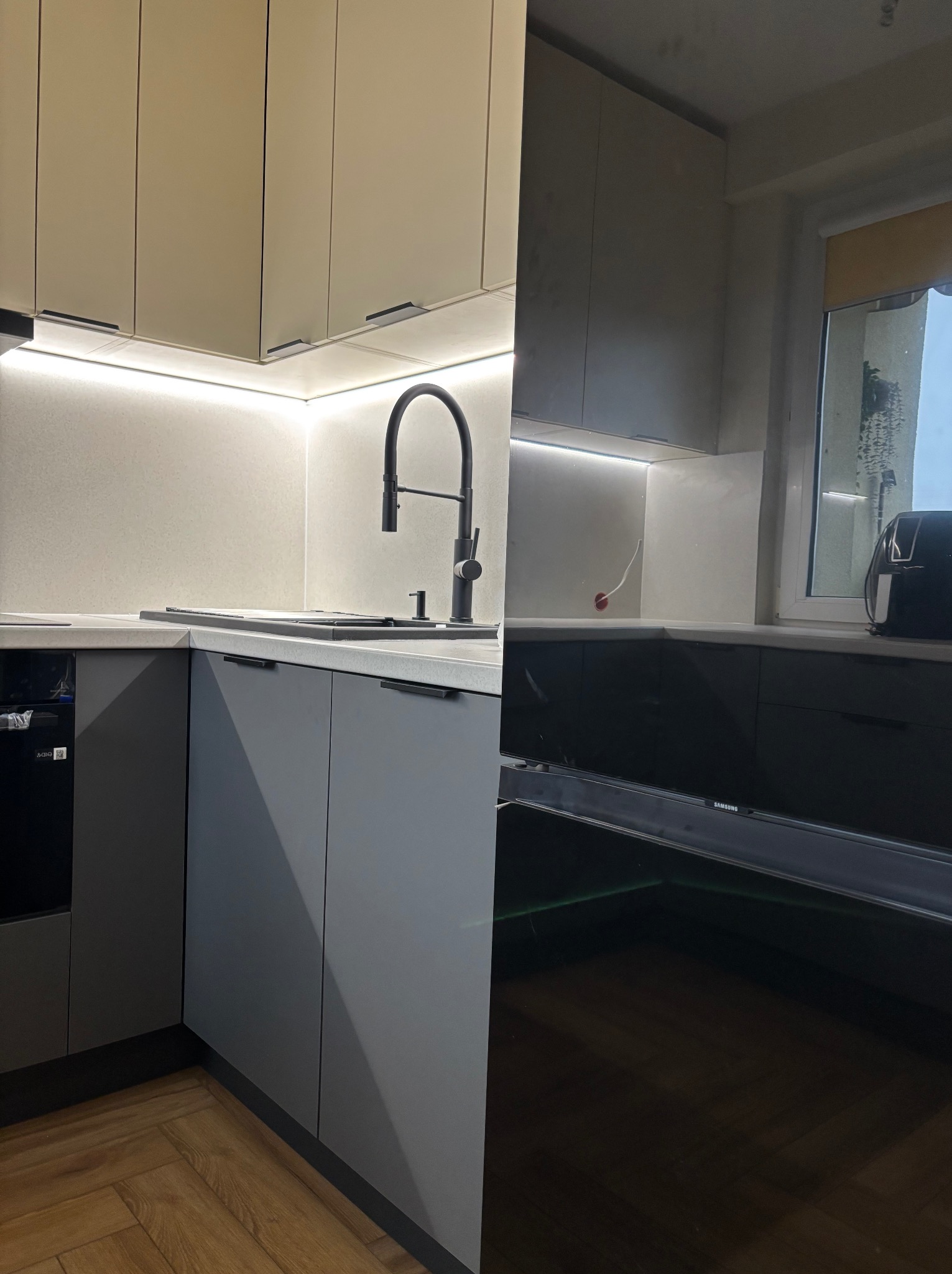 Nowoczesna kuchnia: szare szafki dolne z czarnymi uchwytami, beżowe szafki górne z podświetleniem LED, czarna bateria kuchenna, drewniana podłoga.