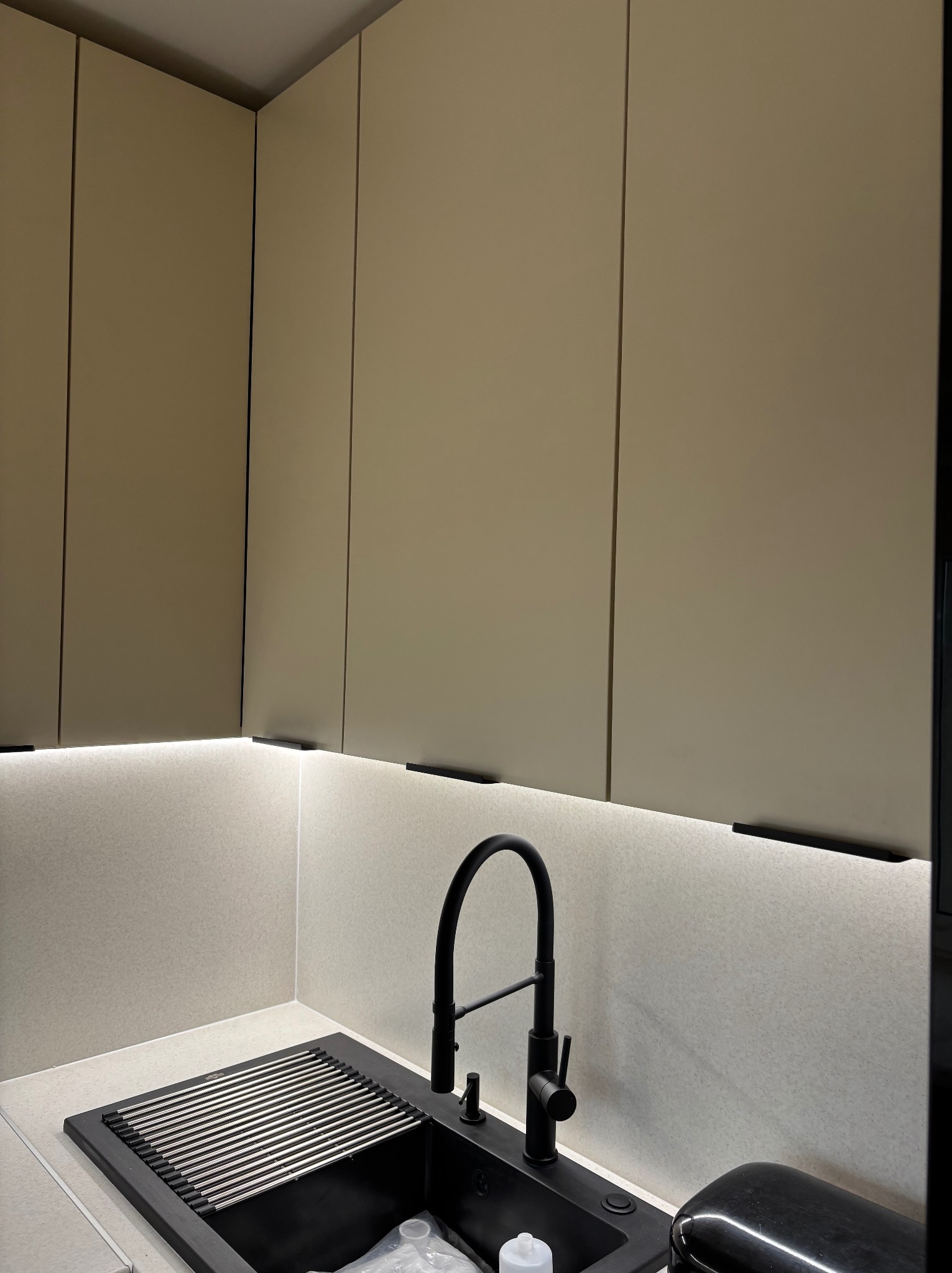 Nowoczesna kuchnia: beżowe szafki wiszące z czarnymi uchwytami, podświetlenie LED pod szafkami, czarny zlew z ociekaczem i baterią, fragment ekspresu do kawy.