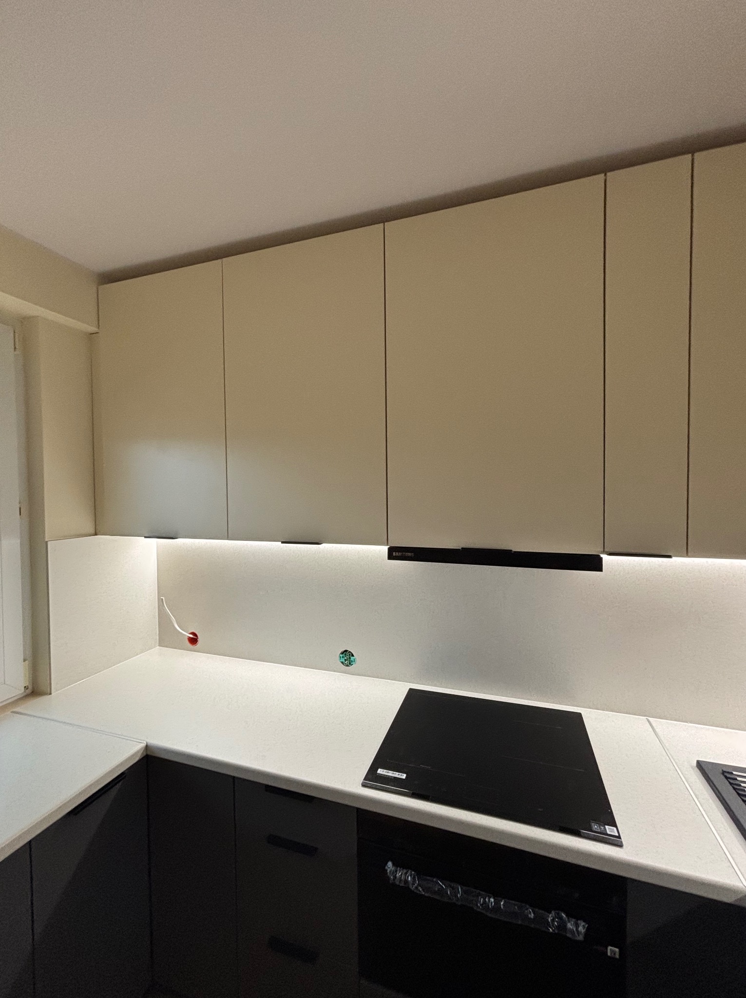 Beżowe szafki kuchenne podświetlone listwą LED, czarny blat z płytą indukcyjną, widoczne gniazdko elektryczne i niedokończone podłączenie kabla.