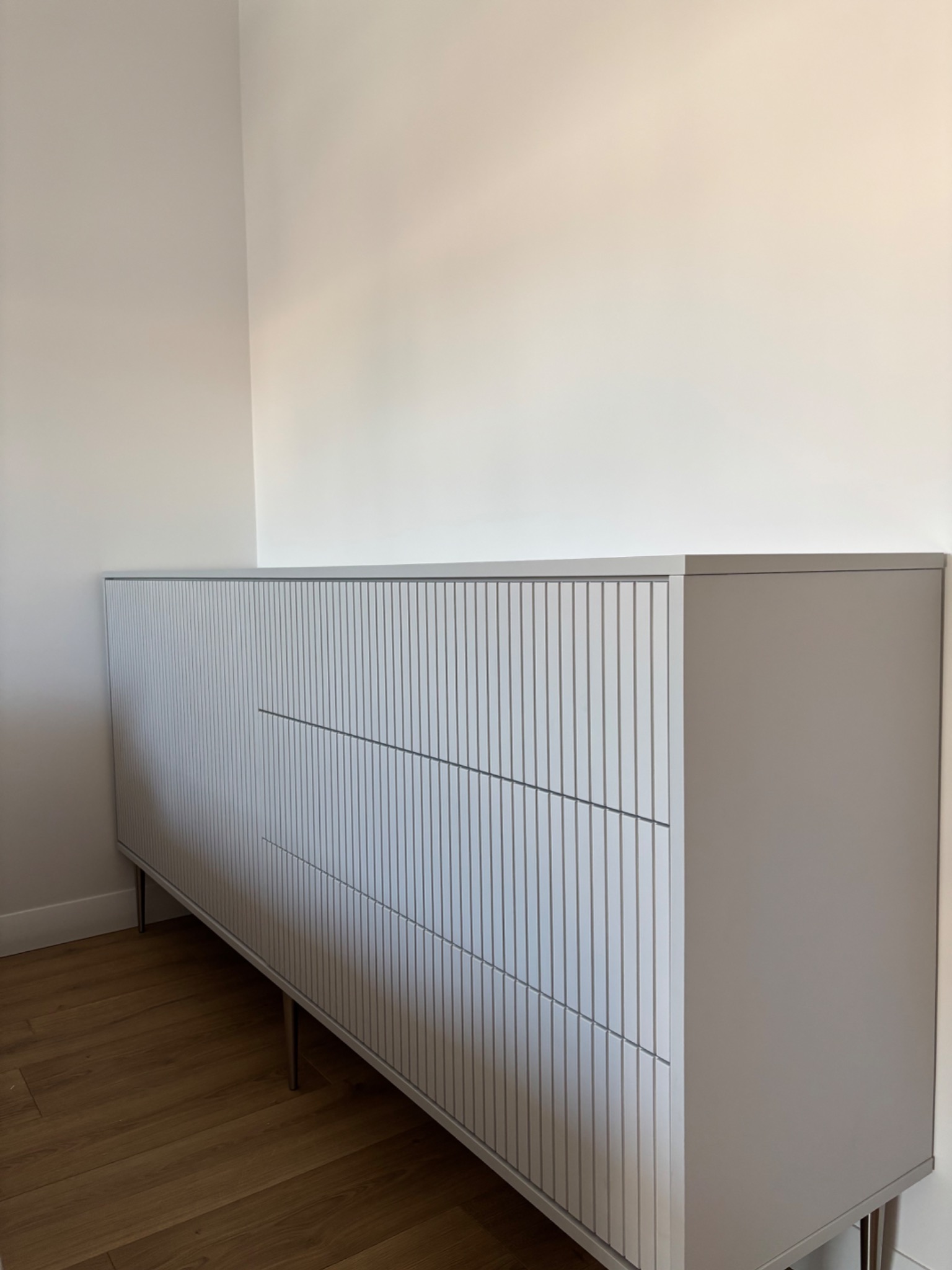 Nowoczesna, długa komoda w odcieniach szarości z ryflowanymi frontami, osadzona na metalowych nóżkach. Minimalistyczny design, jasne wnętrze.