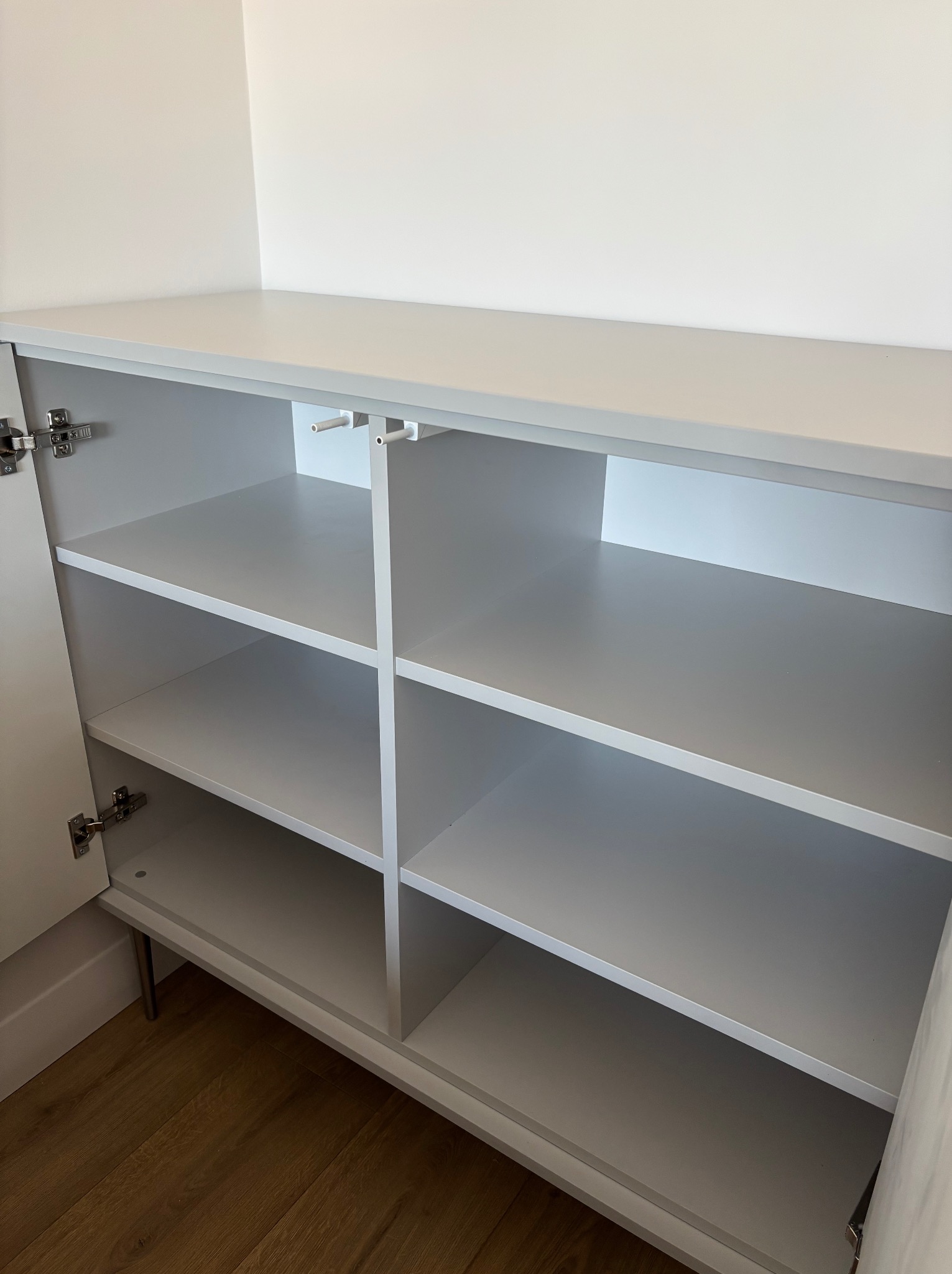 Wnętrze szafki z półkami w odcieniach szarości, widoczne zawiasy i fragment podłogi z paneli. Minimalistyczny design, proste linie.