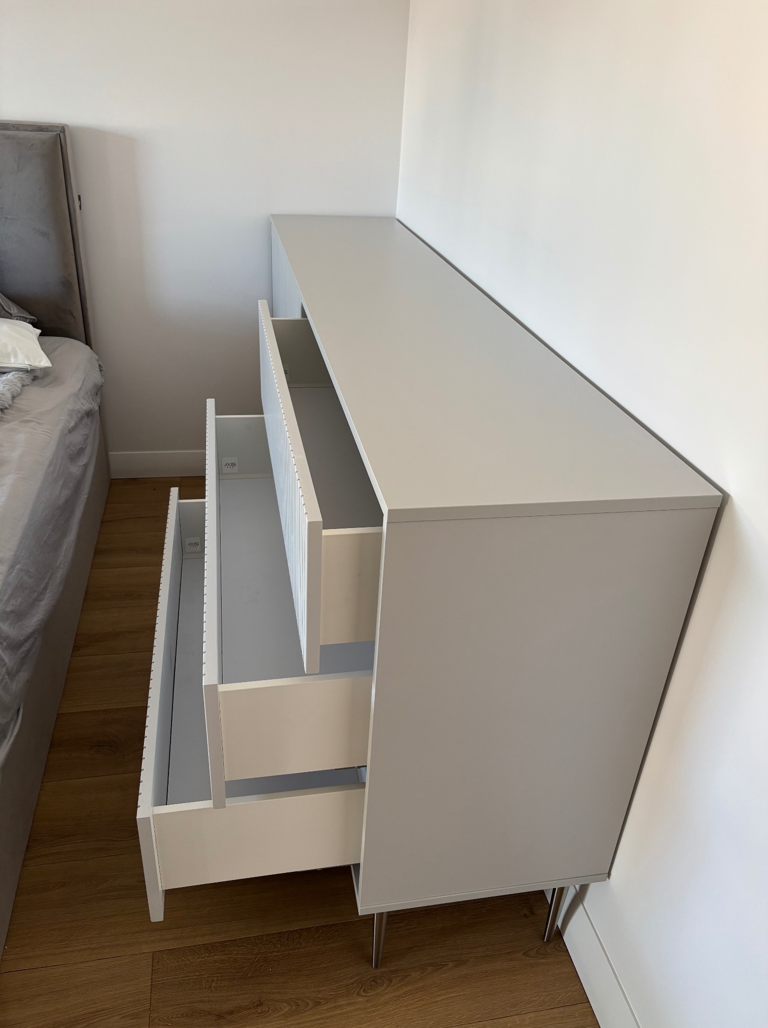 Szara komoda z trzema wysuniętymi szufladami na metalowych nóżkach, widoczna obok łóżka na drewnianej podłodze, na tle białej ściany.