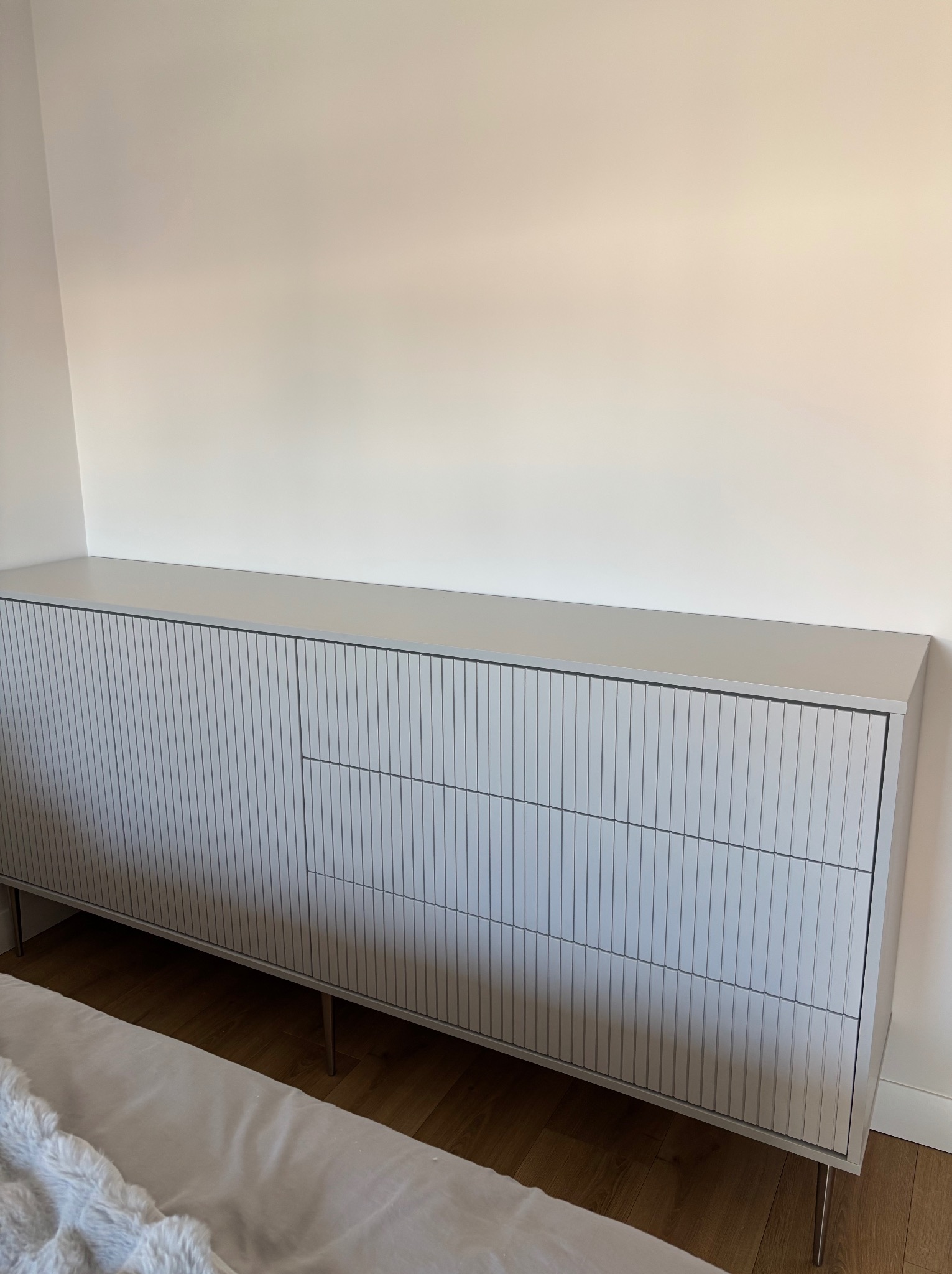 Jasnoszara komoda na metalowych nóżkach z ryflowanymi frontami szuflad, stojąca na drewnianej podłodze przy białej ścianie w sypialni.