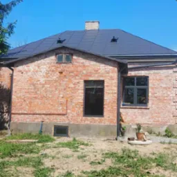 Ceglany dom jednorodzinny z nowym, ciemnoszarym dachem, widok z zewnątrz w słoneczny dzień. Na pierwszym planie trawa i ziemia, w tle błękitne niebo.