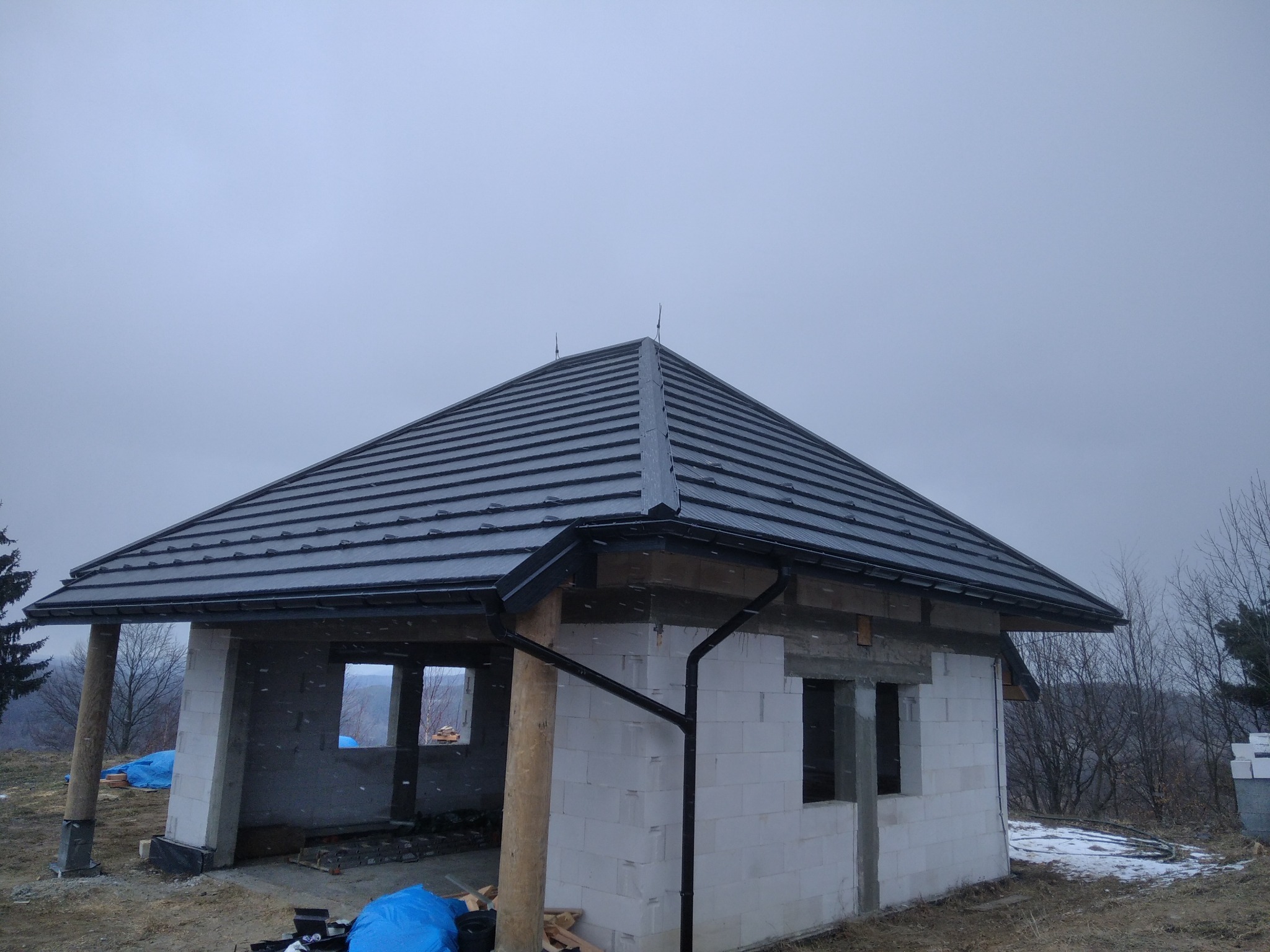 Surowy stan budynku z szarą dachówką, widoczne obróbki blacharskie i rynny, w tle zalesione wzgórza pod pochmurnym niebem.