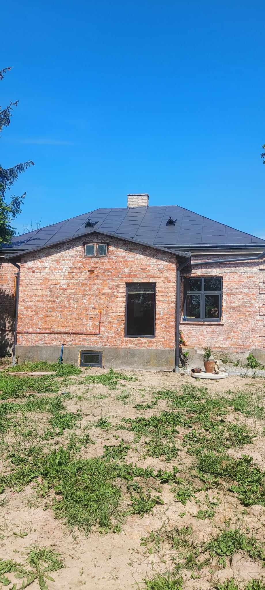 Ceglany dom jednorodzinny z nowym, ciemnoszarym dachem, widok z zewnątrz w słoneczny dzień. Na pierwszym planie trawa i ziemia, w tle błękitne niebo.