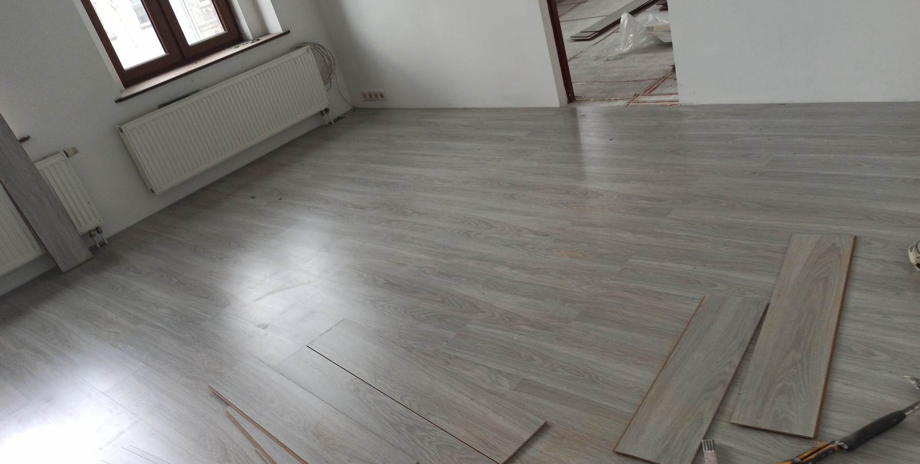 Układanie jasnoszarych paneli podłogowych w pokoju z białymi ścianami i grzejnikiem, widoczne narzędzia i nieukończona podłoga w przejściu do innego pomieszczenia.
