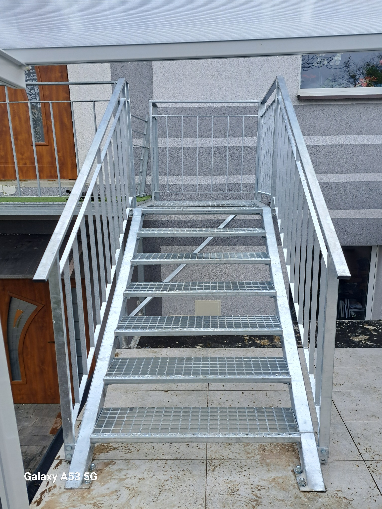 Zewnętrzne, ocynkowane schody metalowe z kratowymi stopniami i balustradą, prowadzące do drzwi balkonowych na tle szarej elewacji budynku.