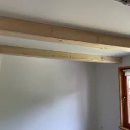 Dwie drewniane belki zamontowane pod sufitem w jasnym pokoju z oknem, widok na zewnątrz.