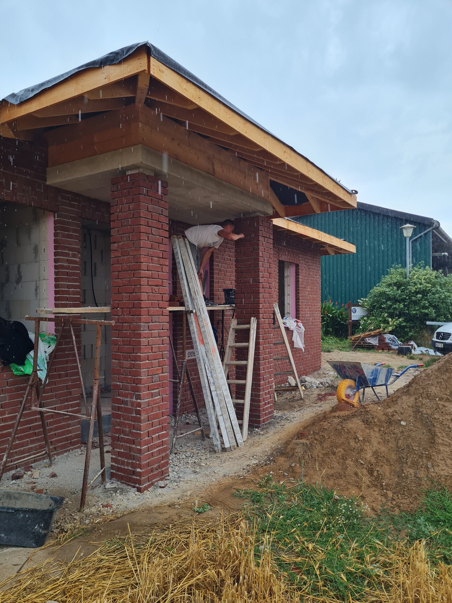 Budowa domu z czerwonej cegły w trakcie deszczu; widoczne rusztowania, drabiny, materiały budowlane i pracownik przy drewnianej konstrukcji dachu.