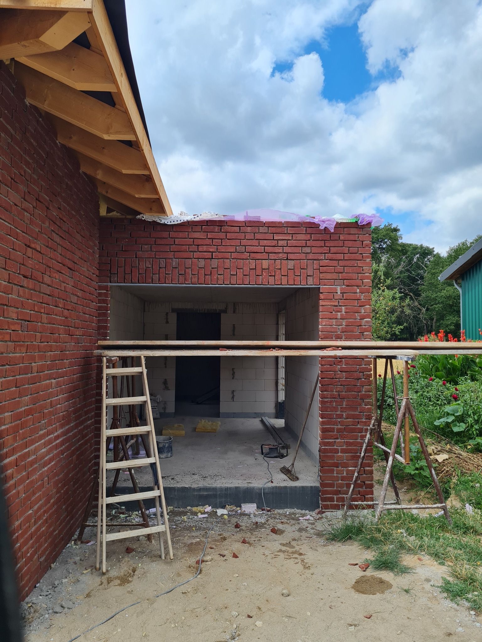 Wznoszenie ścian z czerwonej cegły, widoczne rusztowanie, drabina i fragment dachu z drewnianą konstrukcją, w tle niebieskie niebo z chmurami i zieleń
