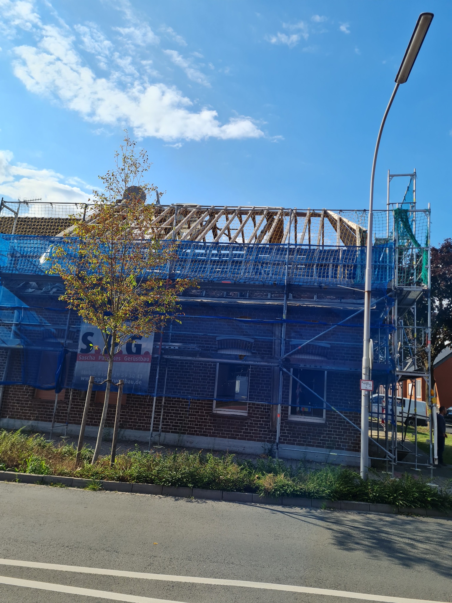 Budynek z cegły w trakcie remontu dachu, otoczony rusztowaniem z niebieską siatką zabezpieczającą, widoczny szkielet dachu i błękitne niebo.