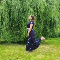 Młoda kobieta w dwuczęściowej, granatowej sukience w białe kwiaty, pozuje na tle płaczącej wierzby, unosząc jedną nogę w górę.