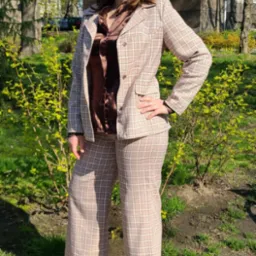 Kobieta w brązowym, kraciastym garniturze (szerokie spodnie i marynarka) i satynowej koszuli pozuje na zewnątrz, na tle zieleni i budynku.