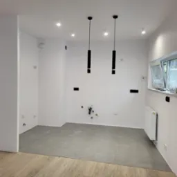 Wnętrze w trakcie prac wykończeniowych: świeżo pomalowane białe ściany, szara podłoga z płytek, widoczne instalacje hydrauliczne i elektryczne, nowoczesne czarne lampy wiszące, grzejnik pod oknem.