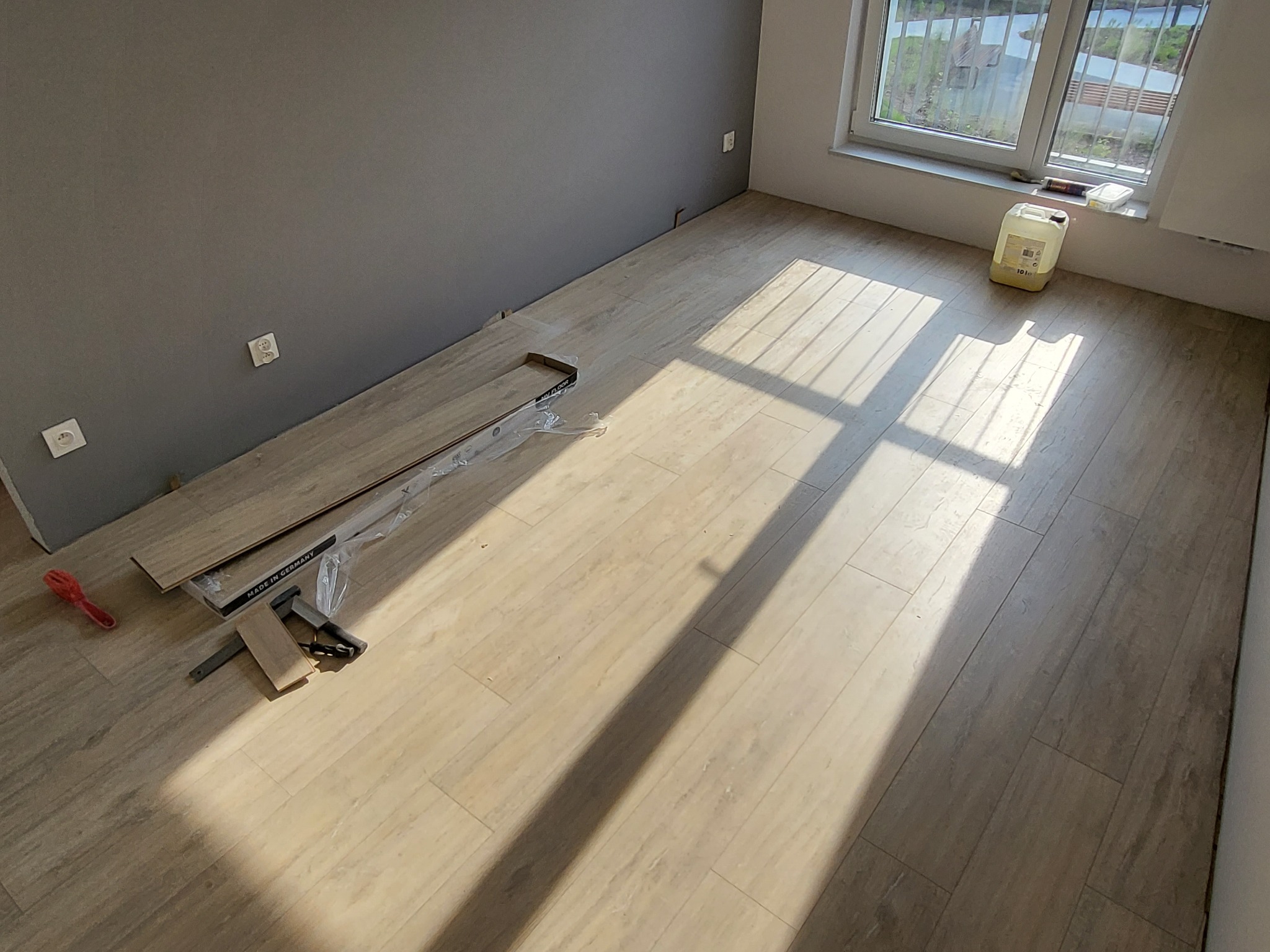 Pokój w trakcie prac wykończeniowych: częściowo ułożona podłoga z paneli, paczka paneli pod ścianą, narzędzia montażowe i kanister z płynem przy oknie.