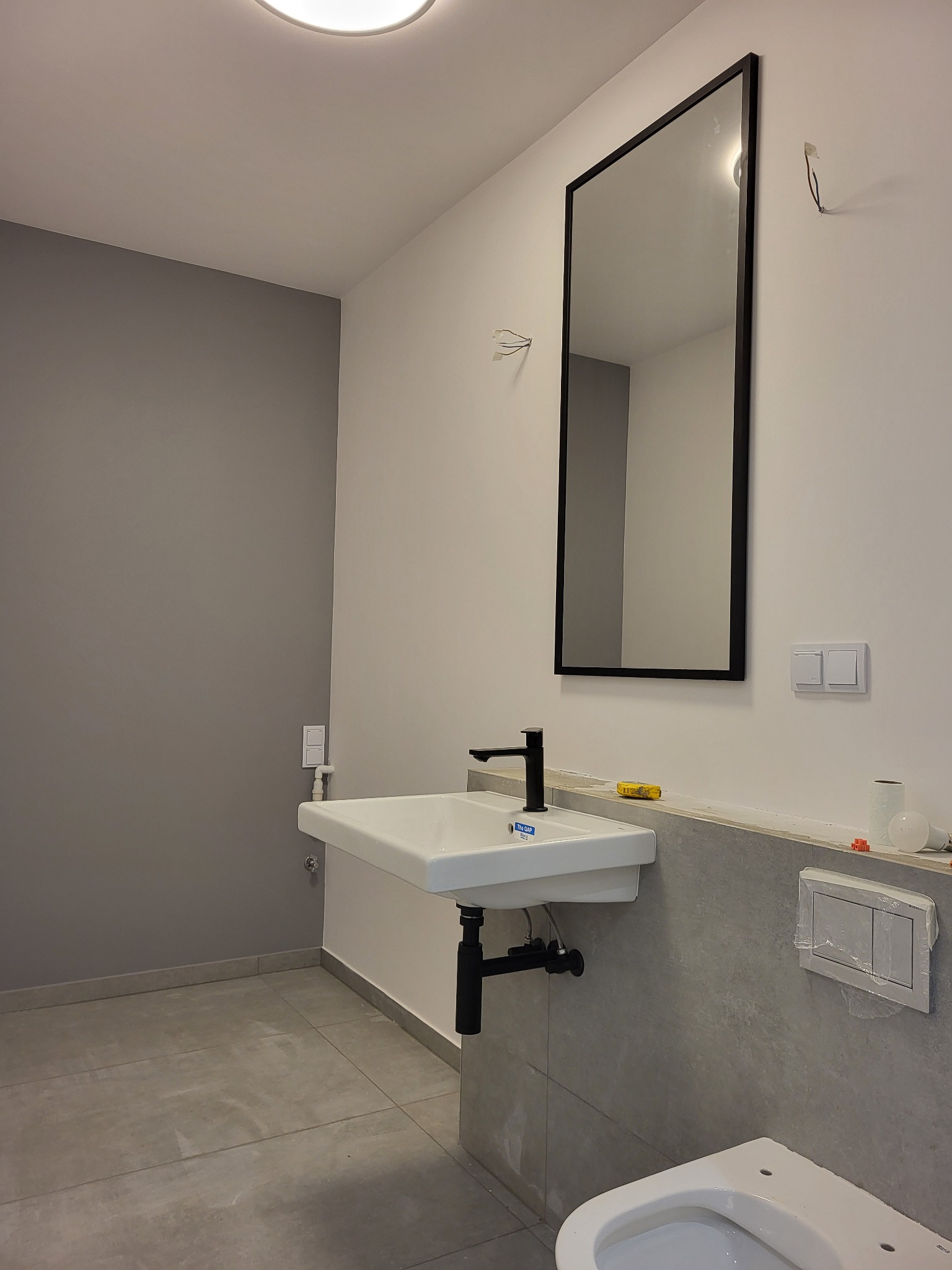 Nowoczesna łazienka w trakcie wykończenia: biała umywalka z czarną armaturą, duże lustro w czarnej ramie, szare płytki na ścianie, widoczne elementy instalacji i narzędzia, surowy stan wykończenia.