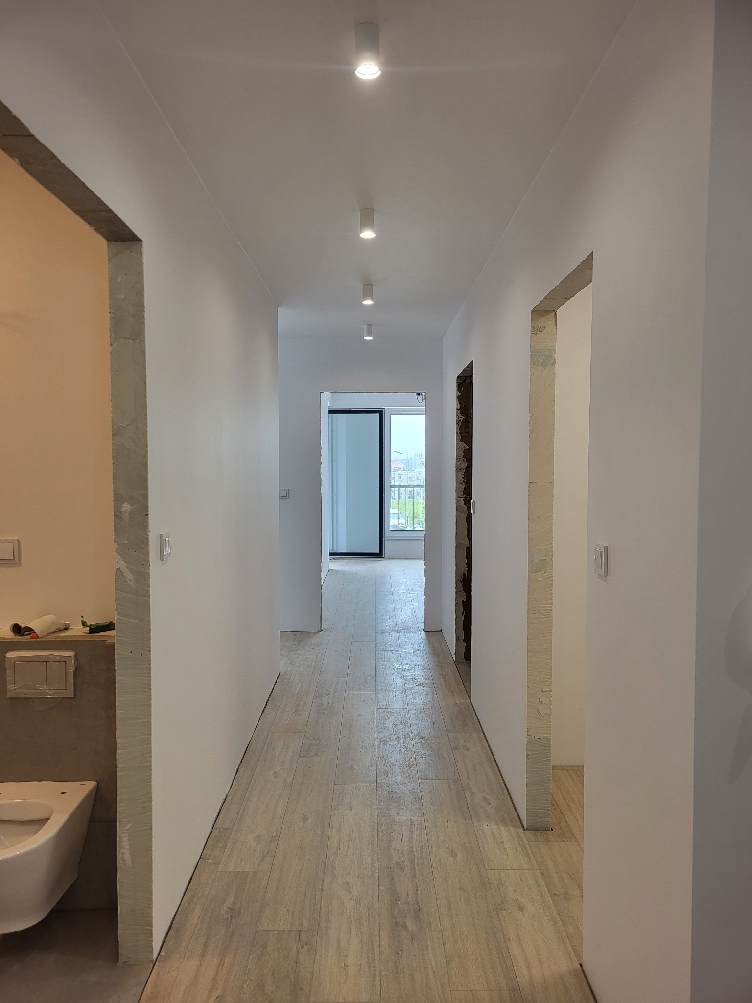 Korytarz w trakcie wykańczania, z widoczną podłogą z paneli imitujących drewno, świeżo pomalowanymi ścianami na biało i otworami drzwiowymi w stanie surowym.