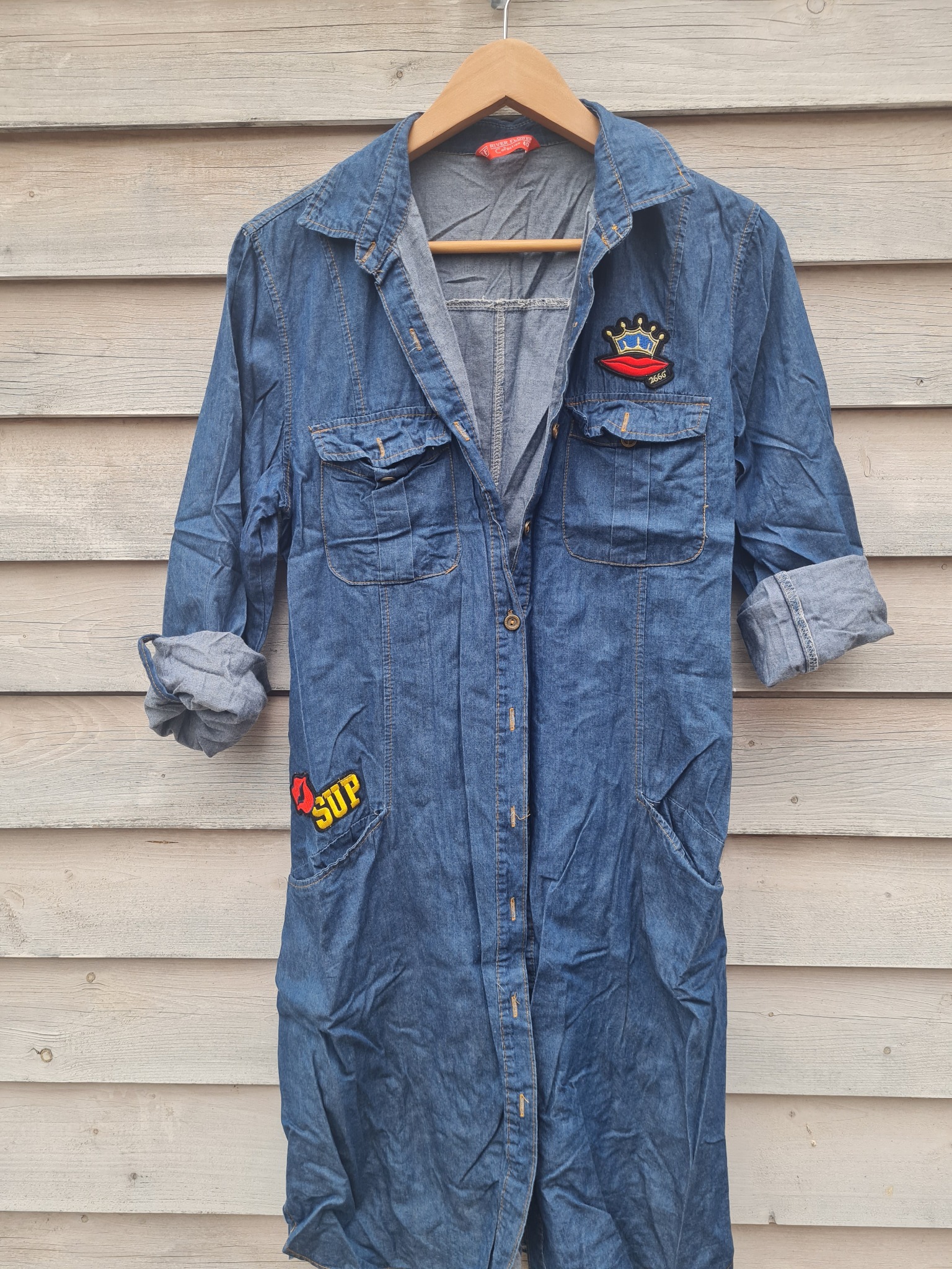 Dżinsowa sukienka koszulowa z podwiniętymi rękawami, wisząca na drewnianym wieszaku na tle jasnych desek, ozdobiona naszywkami z motywami ust i korony.