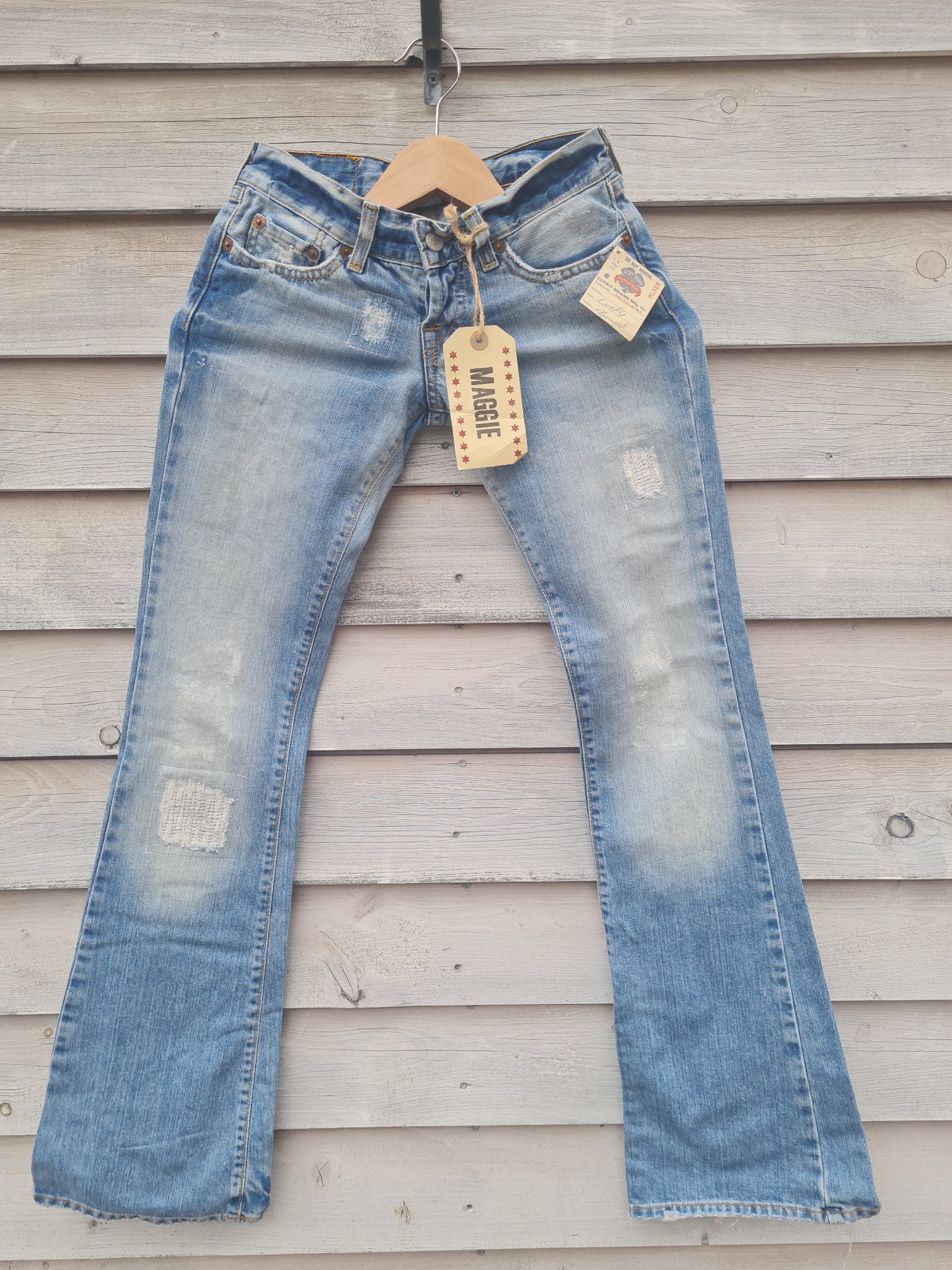 Jasne dżinsy damskie typu bootcut z przetarciami i naszywką na kolanie, wiszące na drewnianym wieszaku przed szarą, drewnianą ścianą, z etykietą marki Maggie.