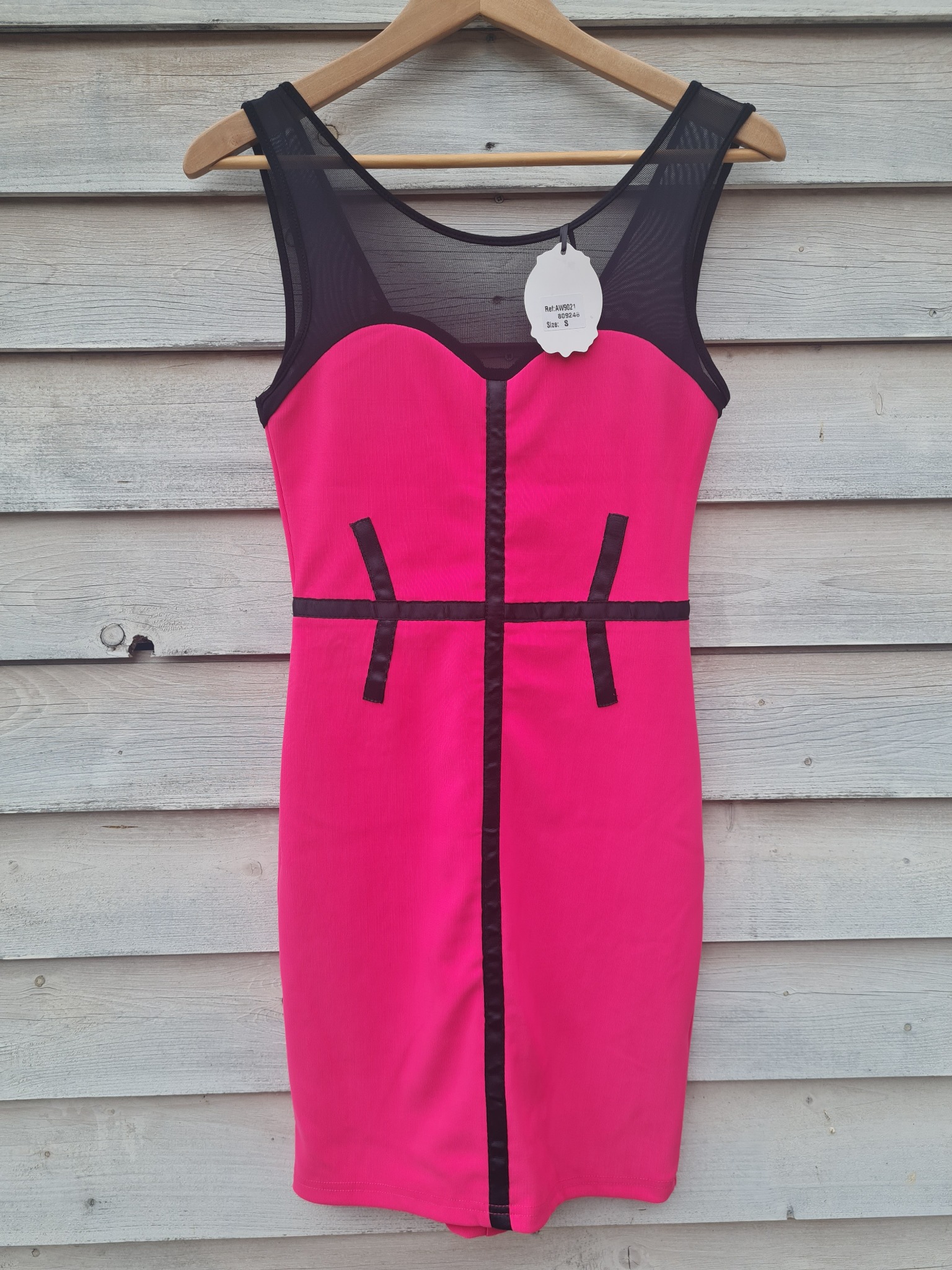 Różowa, dopasowana sukienka z czarnymi, geometrycznymi wstawkami, wisząca na drewnianym wieszaku przed szarą, drewnianą ścianą.