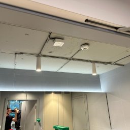 RUSLAN IEGER FPC - Szare wnętrze z widocznymi przewodami elektrycznymi poprowadzonymi po suficie, cylindryczne lampy wiszące, czujnik dymu oraz odblask w lustrze z widoczną osobą robiącą zdjęcie.