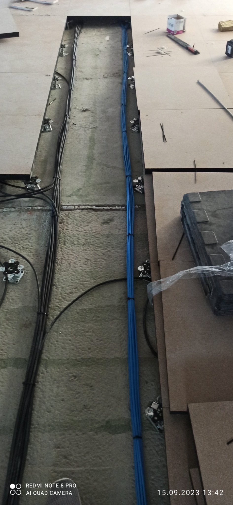 Widok z góry na koryto w podłodze technicznej z ułożonymi kablami: czarnymi po lewej i niebieskimi po prawej stronie, zamocowanymi uchwytami do podłoża. Fragmenty płyt podłogowych obok koryta...