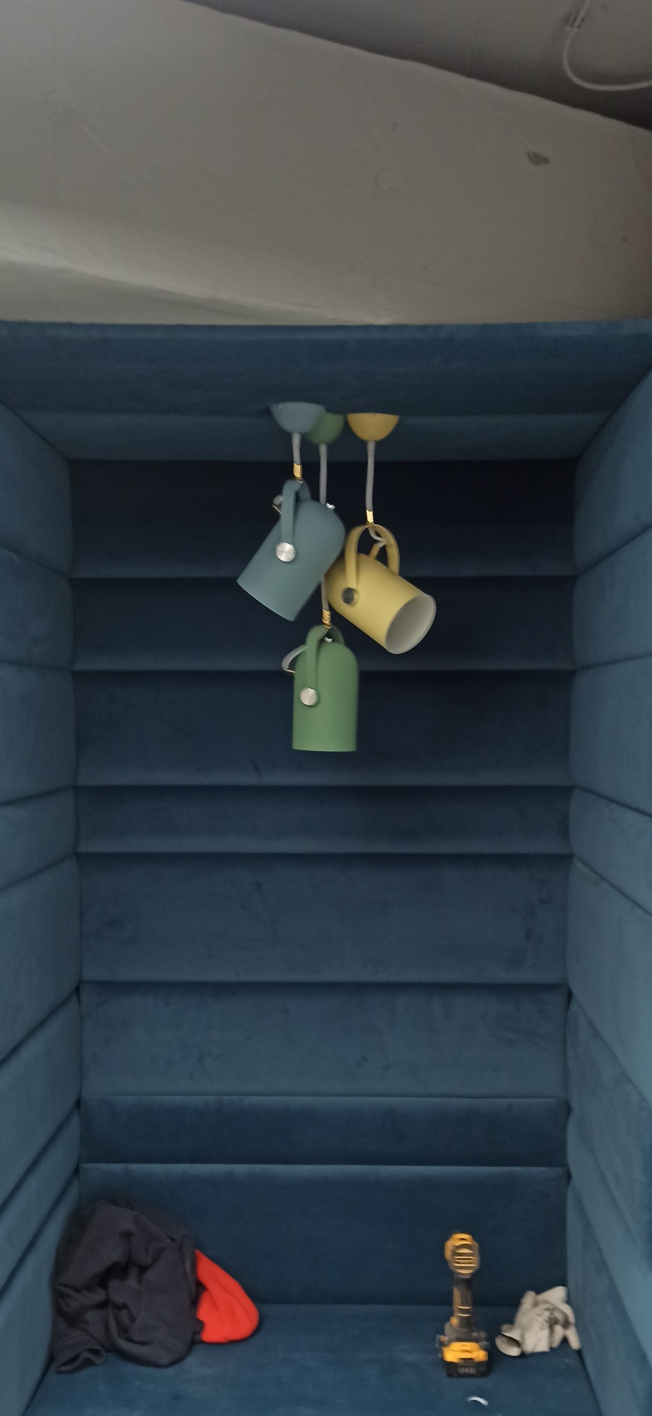 Nietypowa instalacja oświetleniowa z trzema lampami w kształcie kubków w kolorach pastelowych: niebieskim, żółtym i zielonym, zwisająca z sufitu w małym pokoju wyłożonym niebieską tapicerką;...