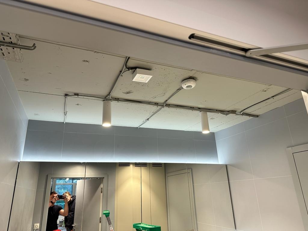 Szare wnętrze z widocznymi przewodami elektrycznymi poprowadzonymi po suficie, cylindryczne lampy wiszące, czujnik dymu oraz odblask w lustrze z widoczną osobą robiącą zdjęcie.