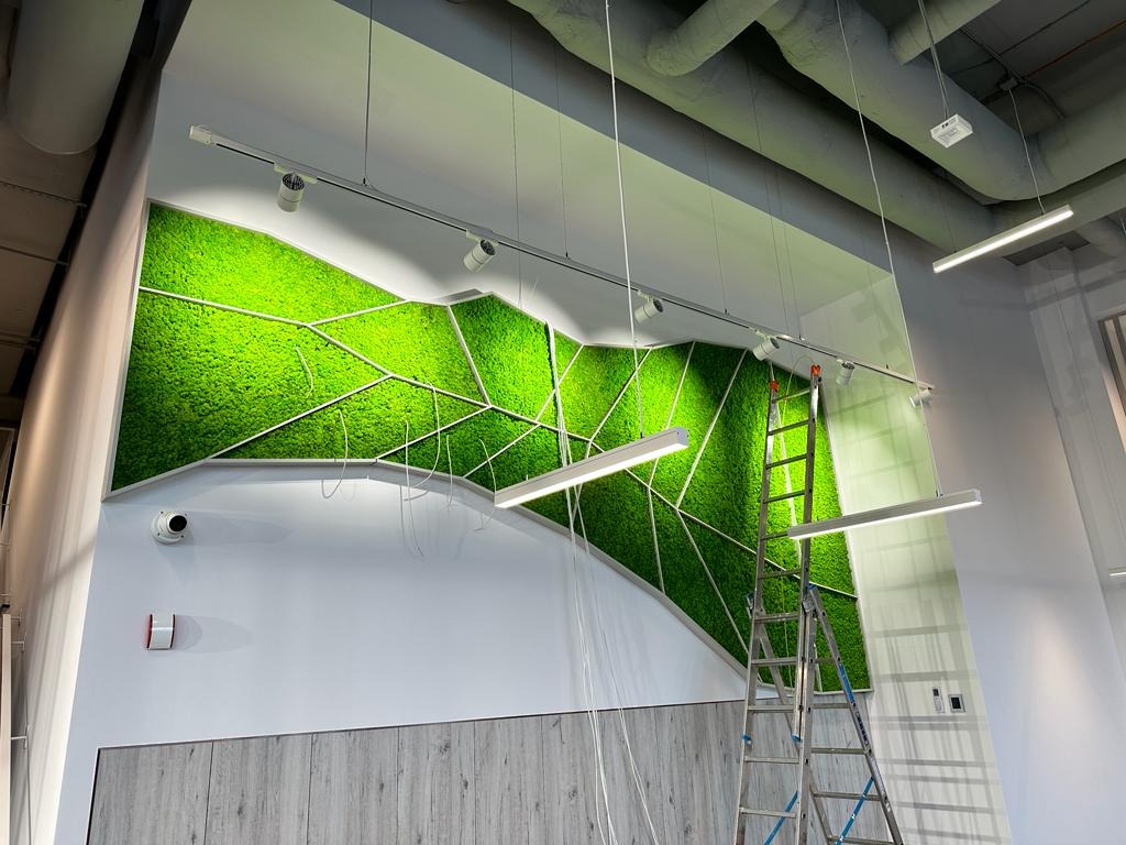 Montaż oświetlenia LED na szynie podsufitowej, z widocznym panelem z mchu dekoracyjnego na ścianie i drabiną. Nowoczesne wnętrze biurowe.