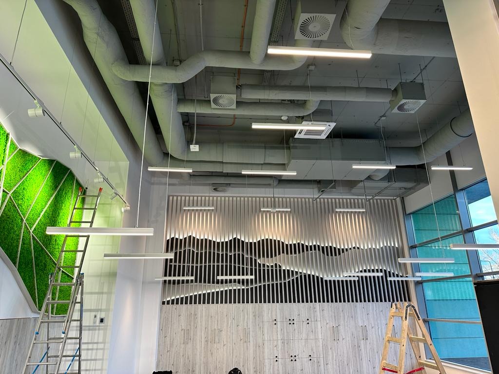 Wnętrze biurowe z zamontowanym oświetleniem liniowym, widoczne drabiny, instalacje wentylacyjne na suficie, dekoracyjna ściana z motywem górskim i panel z mchu.