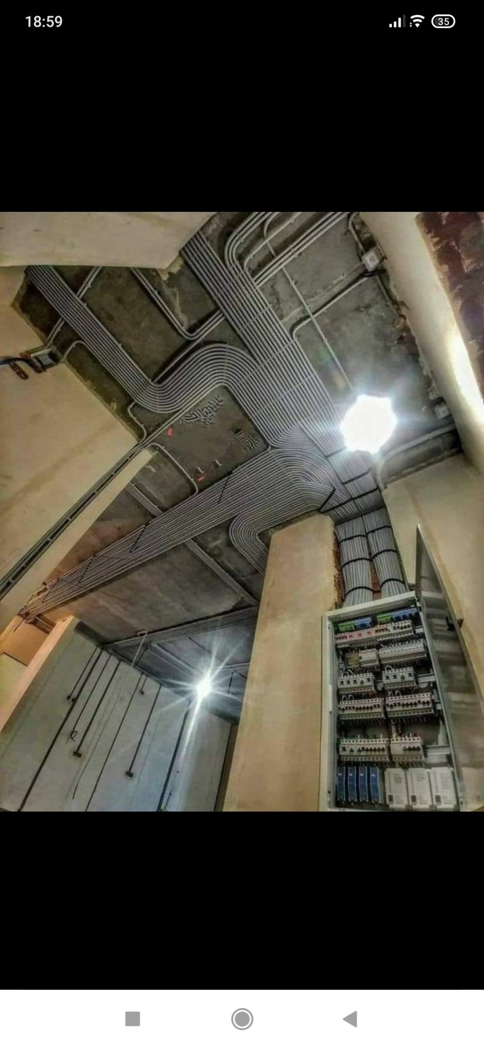 Sufit z widocznymi rurami instalacyjnymi ułożonymi równolegle i rozdzielnicą elektryczną z otwartymi drzwiczkami w surowym stanie budynku.
