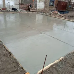 Wylany świeży beton na przygotowanym podłożu pod taras, widoczne drewniane obramowanie i dylatacje, w tle ceglany budynek w trakcie renowacji.