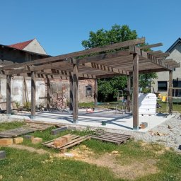 Piotrogród - Montaż drewnianej pergoli z belek na betonowej wylewce, widoczne narzędzia i materiały budowlane w otoczeniu trawnika i budynków.