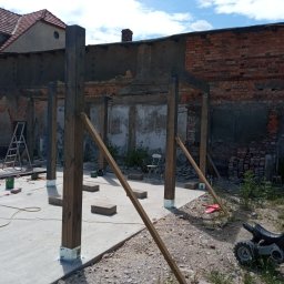 Piotrogród - Konstrukcja drewniana z belek na betonowej wylewce, prawdopodobnie szkielet przyszłej pergoli, z widocznymi metalowymi podstawami słupów, na tle starego budynku z cegły i nieba z chmurami.