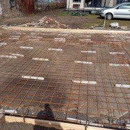 Piotrogród - Przygotowanie podłoża pod taras: siatka zbrojeniowa na betonowej płycie, drewniane obrzeża, widoczne bloki dystansowe, w tle dom z trampoliną i samochód.