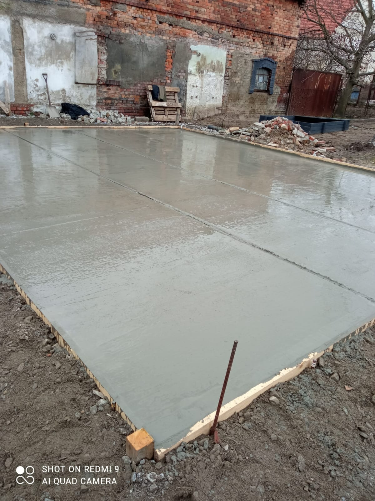 Wylany świeży beton na przygotowanym podłożu pod taras, widoczne drewniane obramowanie i dylatacje, w tle ceglany budynek w trakcie renowacji.