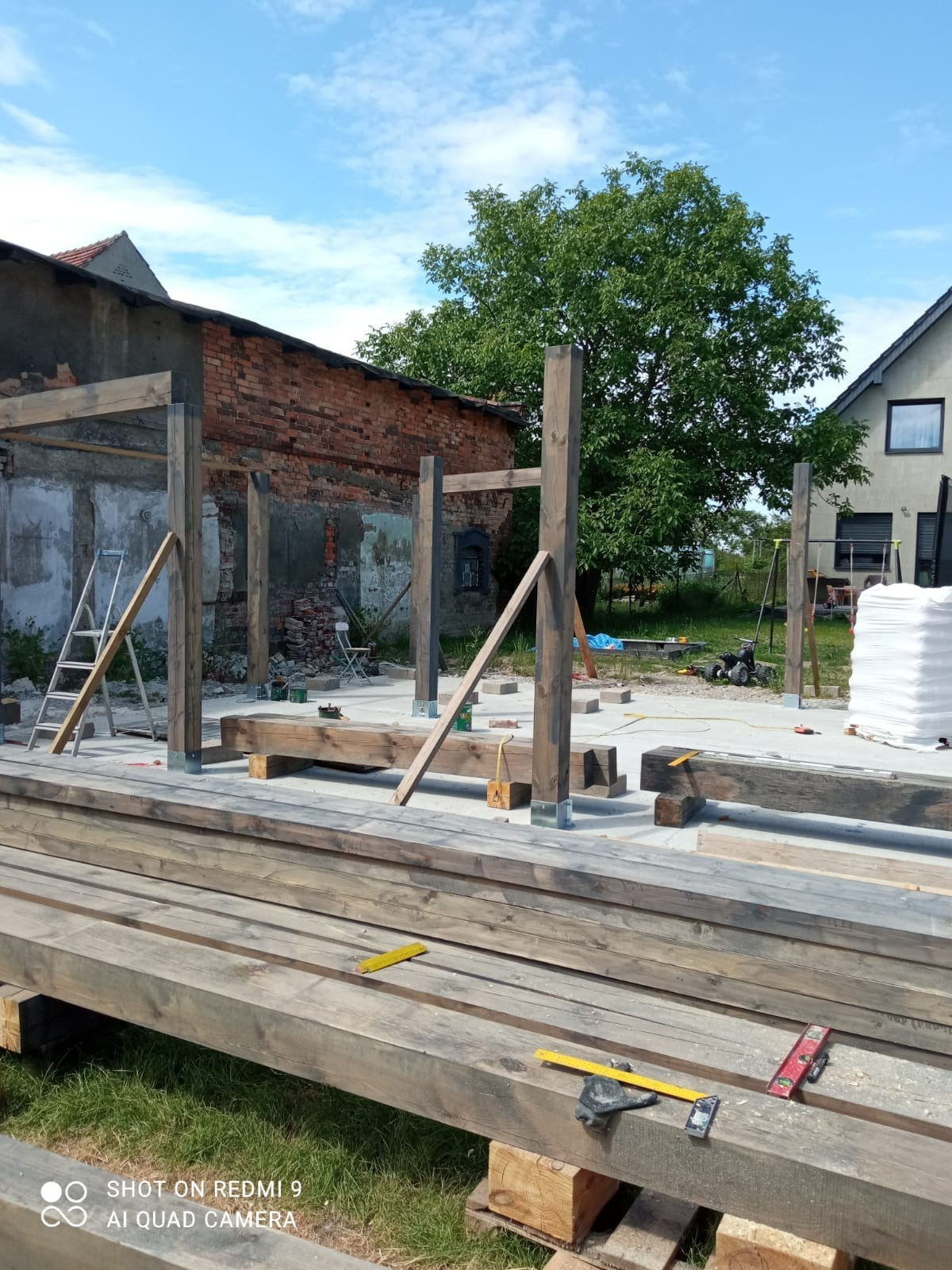 Konstrukcja drewnianej pergoli w trakcie montażu, widoczne słupy i belki, w tle budynek z czerwonej cegły i zieleń drzew