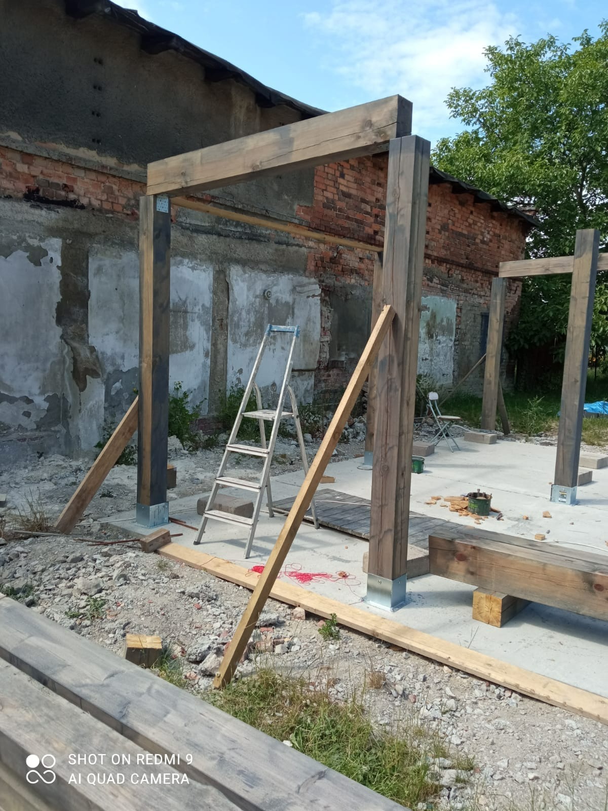 Konstrukcja drewnianej pergoli w trakcie montażu, widoczne słupy i belki zaimpregnowane na szaro, metalowe podstawy słupów, drabina oparta o konstrukcję, w tle budynek z czerwonej cegły.