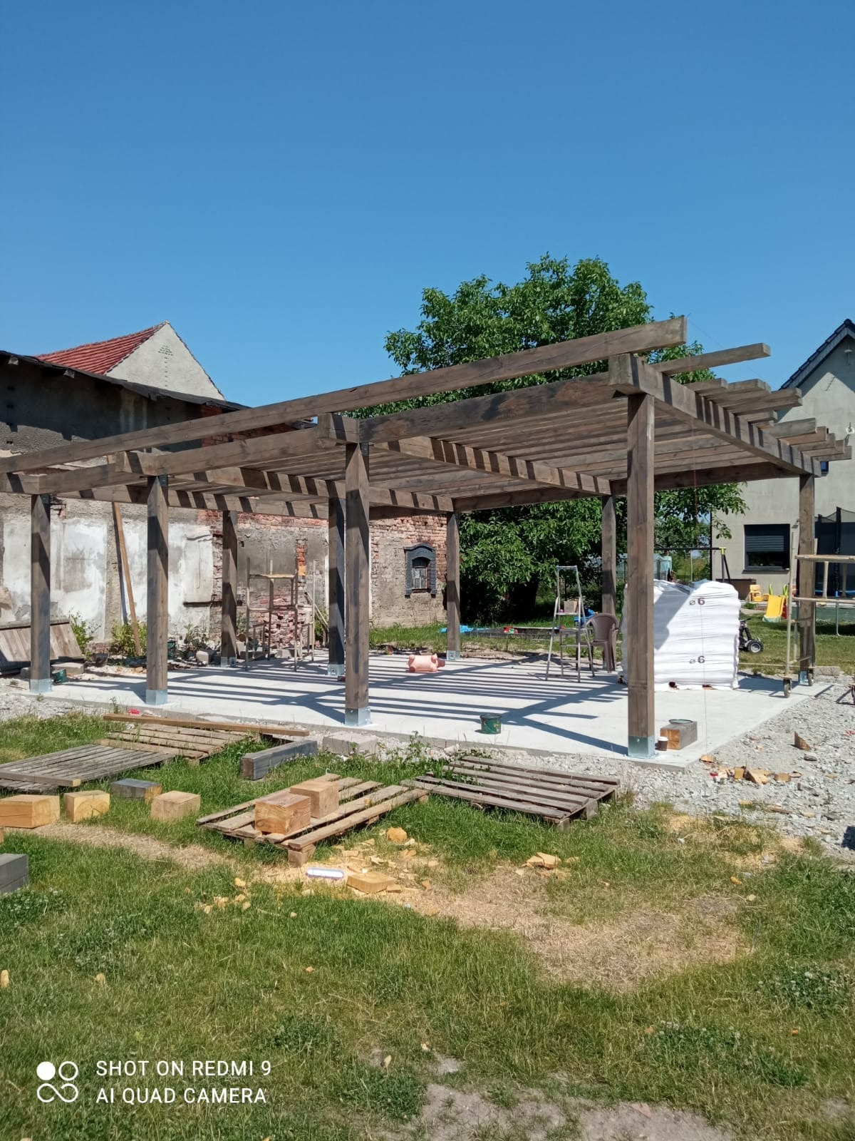 Montaż drewnianej pergoli z belek na betonowej wylewce, widoczne narzędzia i materiały budowlane w otoczeniu trawnika i budynków.