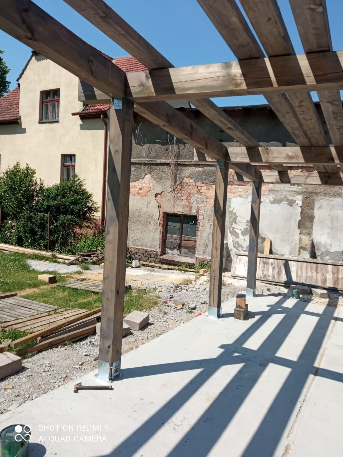 Drewniana pergola w trakcie montażu, widoczne słupy z metalowymi podstawami, betonowa wylewka, w tle budynek mieszkalny i budynek gospodarczy z cegły.